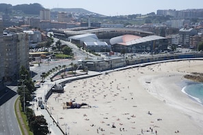 A beach with people sunbathing and swimming, a stadium in the background, and a road running parallel to the beach.