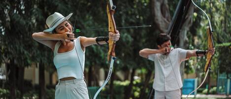 Two individuals practicing archery in an outdoor setting with trees and a bench in the background.