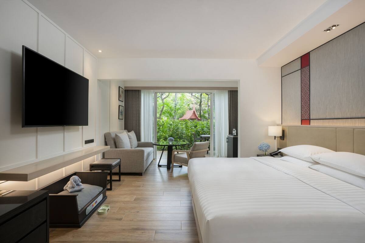 A modern hotel room with a large bed, a flat-screen TV mounted on the wall, a sofa, a small table, and a view of greenery through the window.