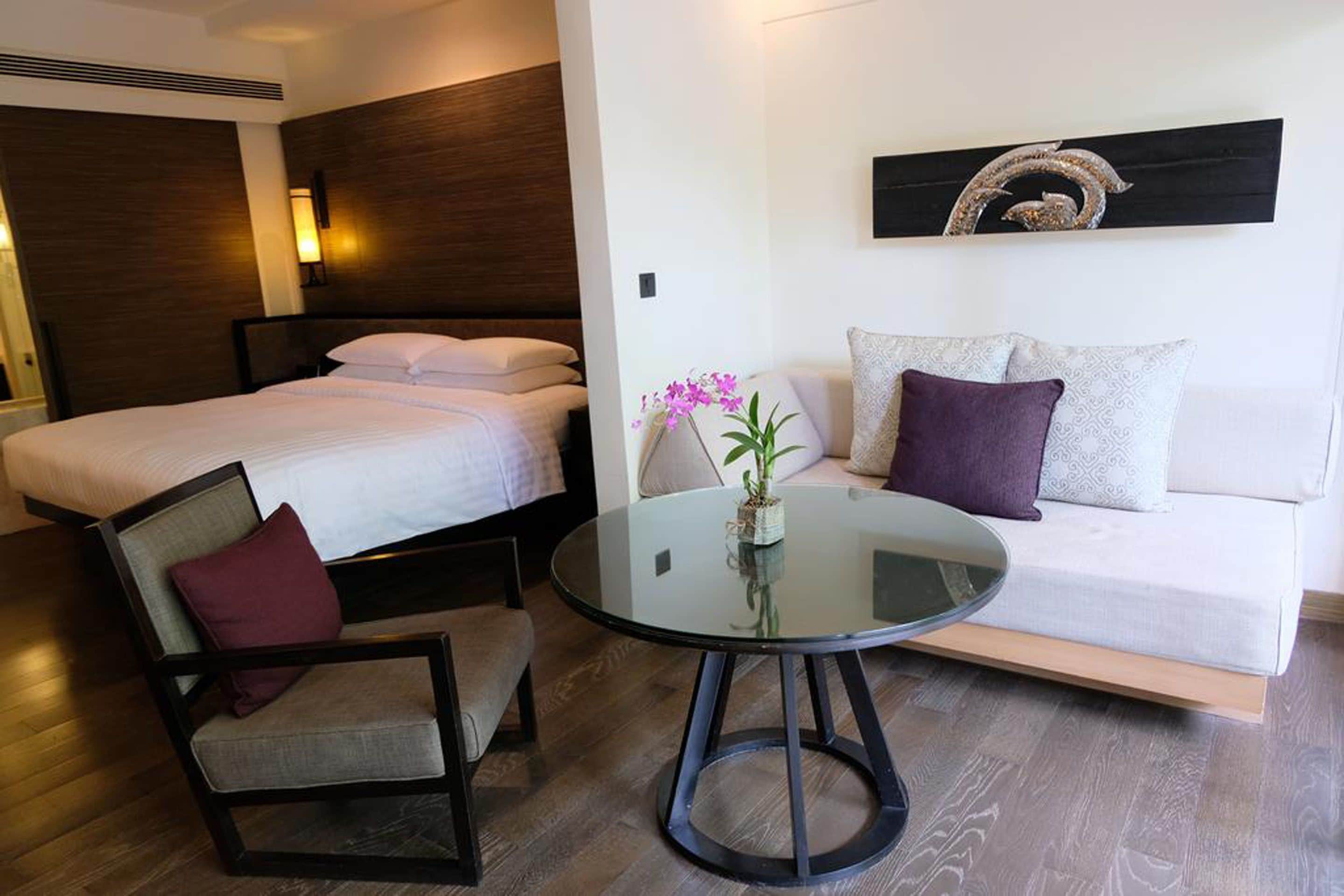 A modern hotel room with a bed, a round glass table, a sofa, and a chair.