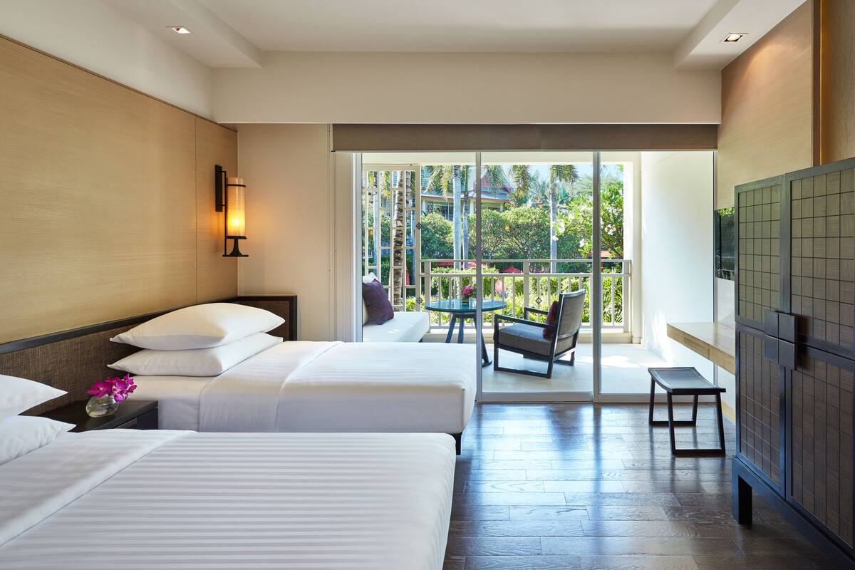 A hotel room with two beds, a small table, a chair, and a view of the outdoors.