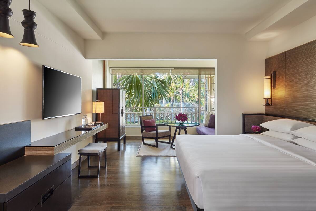 A hotel room with a bed, a desk, a chair, a TV, a balcony with a view of palm trees, and a nightstand with a lamp.