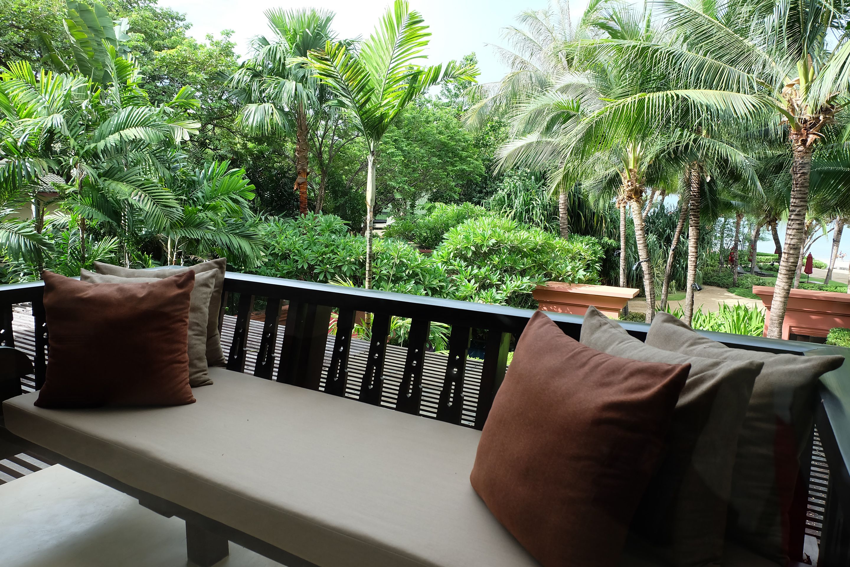 A balcony with a bench and cushions, surrounded by lush greenery and palm trees.