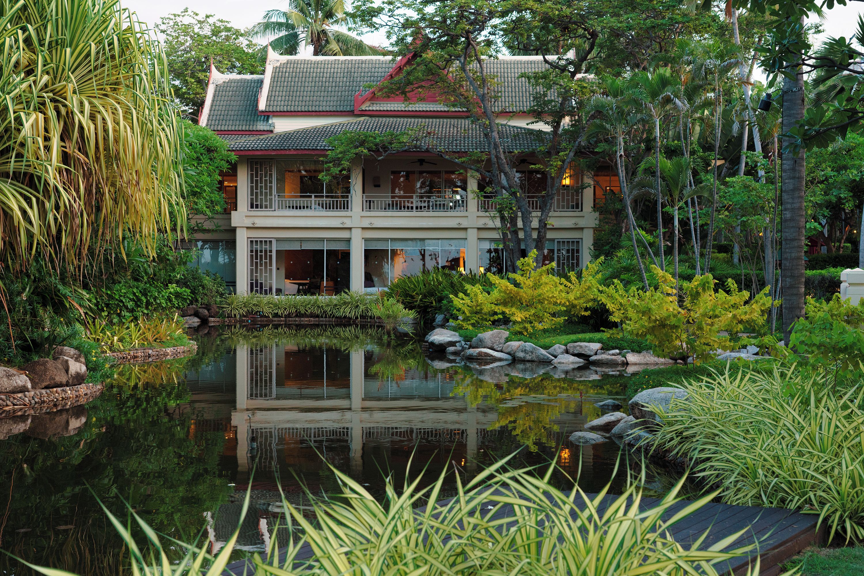 Un bâtiment traditionnel à plusieurs étages, doté d’un toit en tuiles, entouré d’une végétation luxuriante et bordé d’un bassin aux eaux calmes et réfléchissantes.