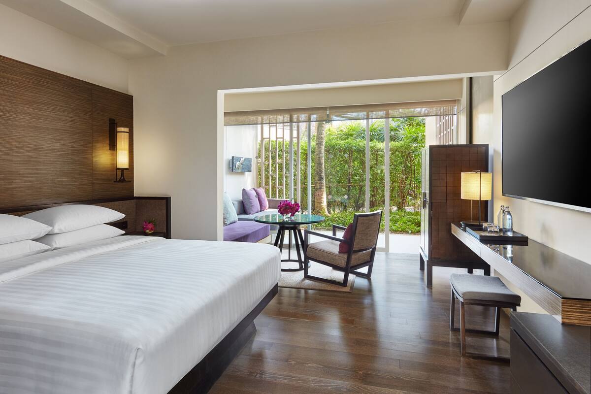 Ein Hotelzimmer mit einem großen Bett, einem Schreibtisch mit Stuhl, einem Fernseher und einem Balkon mit Blick auf Grünflächen.