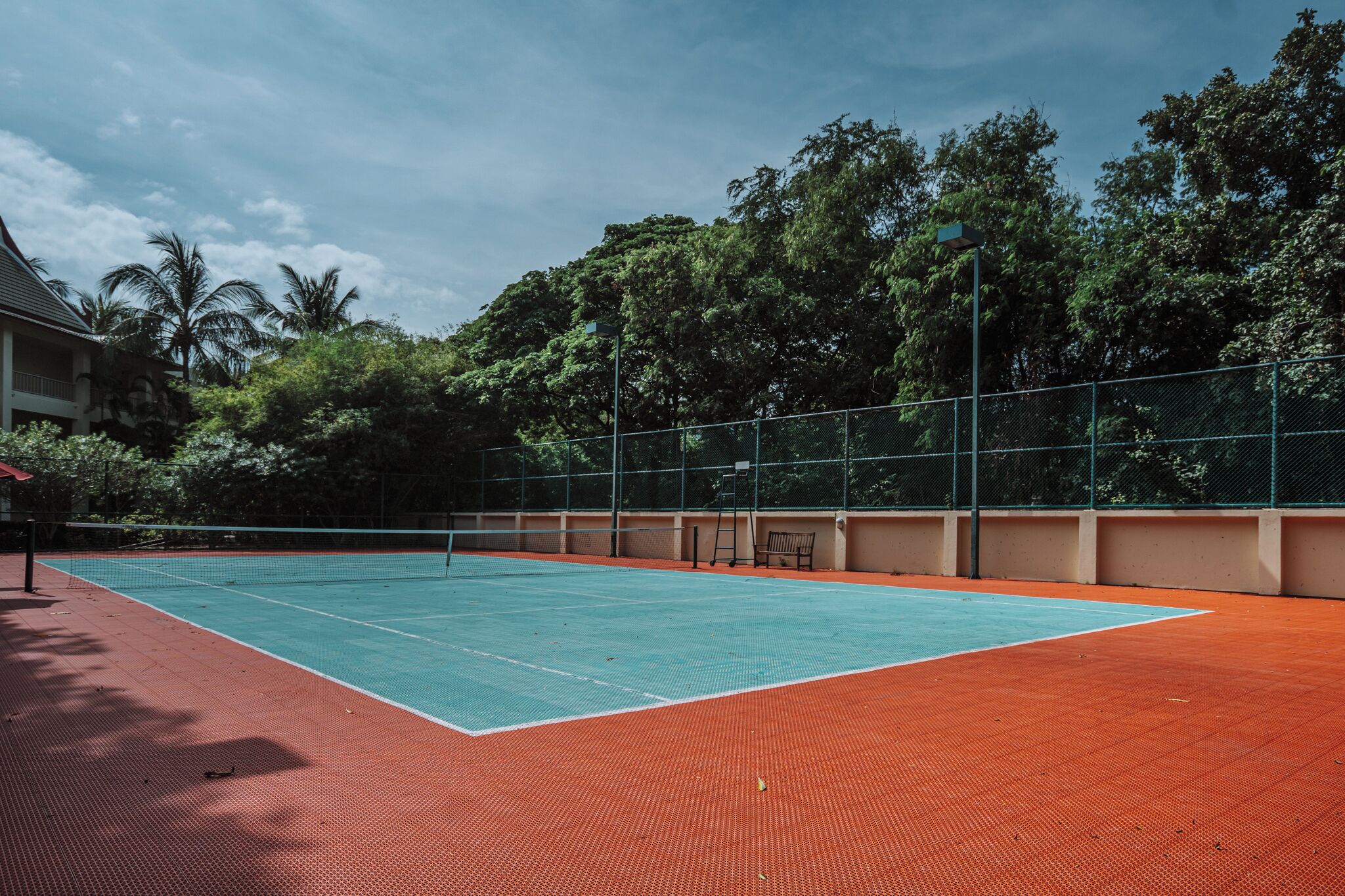 An outdoor tennis court with a net, surrounded by a fence and trees.