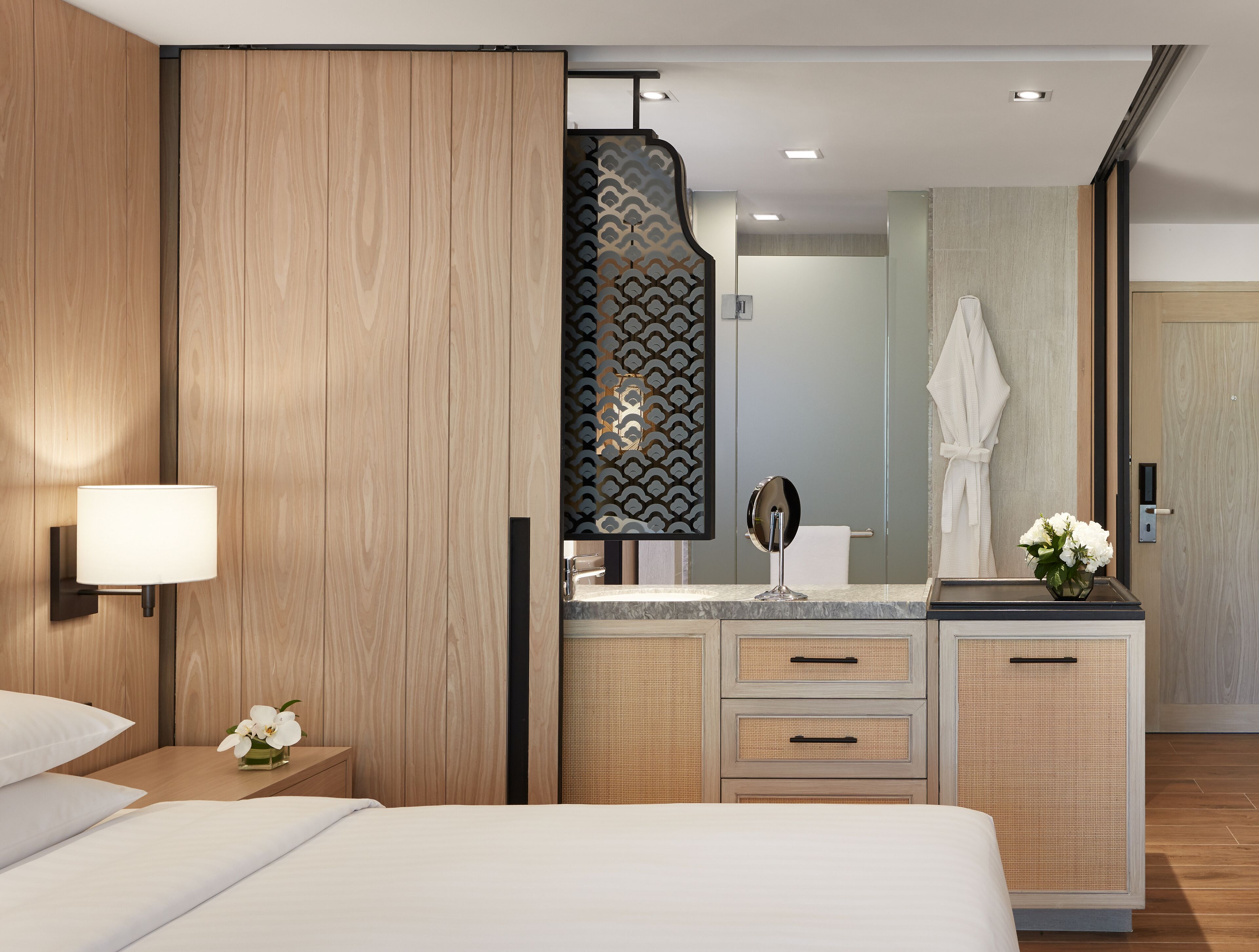 A modern hotel room with a wooden wardrobe, a bedside table with a lamp, a mirror, and a bathroom visible through a glass partition.
