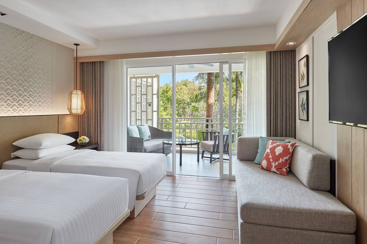 A hotel room with two beds, a sofa, a balcony with a view of greenery, and a flat-screen TV.