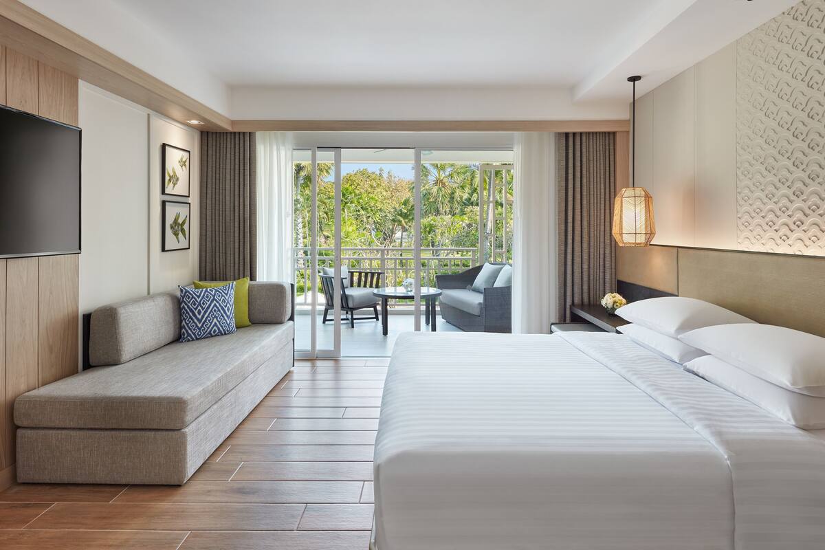 A hotel room with a large bed, a sofa with cushions, a TV, and a balcony with a table and chairs.