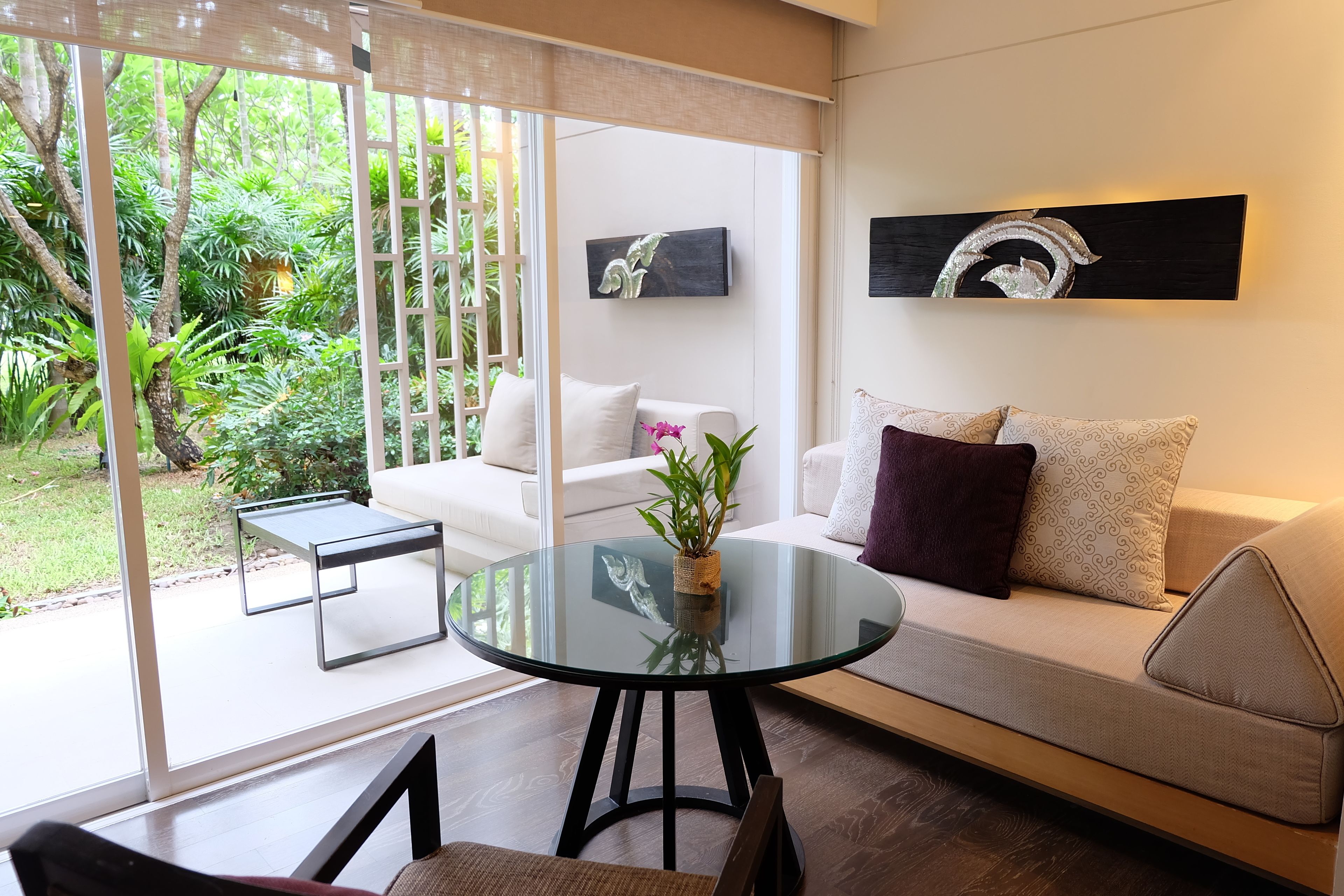 A modern living room with a glass table, a sofa, and a coffee table. There is a painting on the wall and a view of greenery outside.