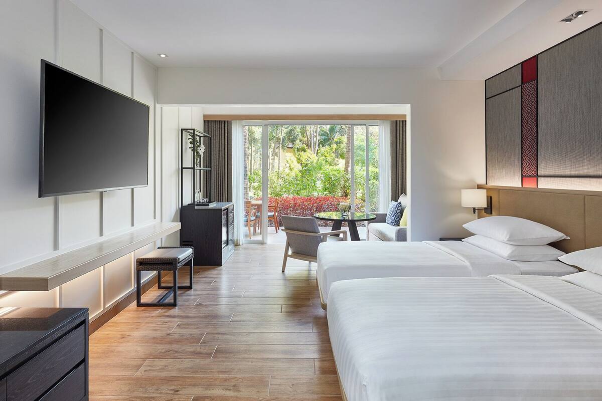 Ein Hotelzimmer mit einem großen Bett, einem an der Wand befestigten Fernseher, einem Schreibtisch mit Stuhl und Blick ins Freie durch eine Glastür.