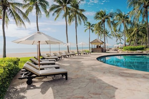 Zona junto a la piscina con sillones de descanso y sombrillas, palmeras y vista al mar.