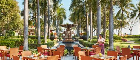 Un restaurante con terraza al aire libre, rodeado de palmeras y una fuente central.