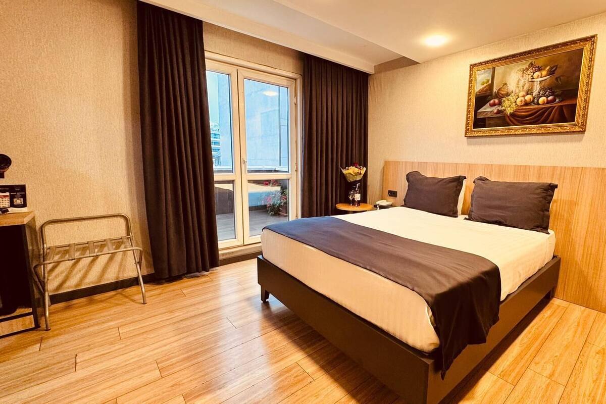 A hotel room with a large bed, a desk with a chair, a painting on the wall, and a view through the window.