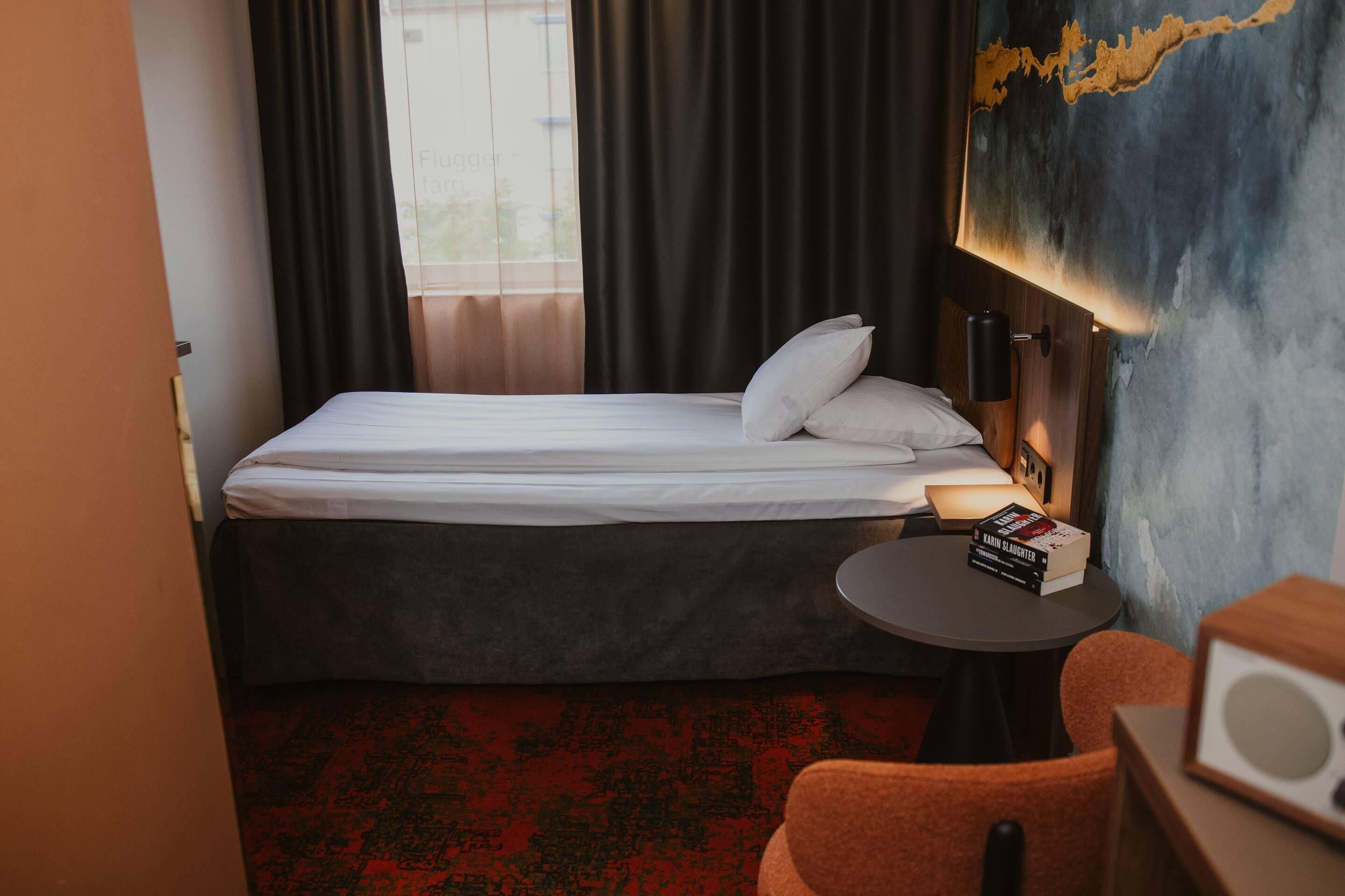 Habitación de hotel con cama, una mesita redonda con libros, una silla y una lámpara fijada a la pared.
