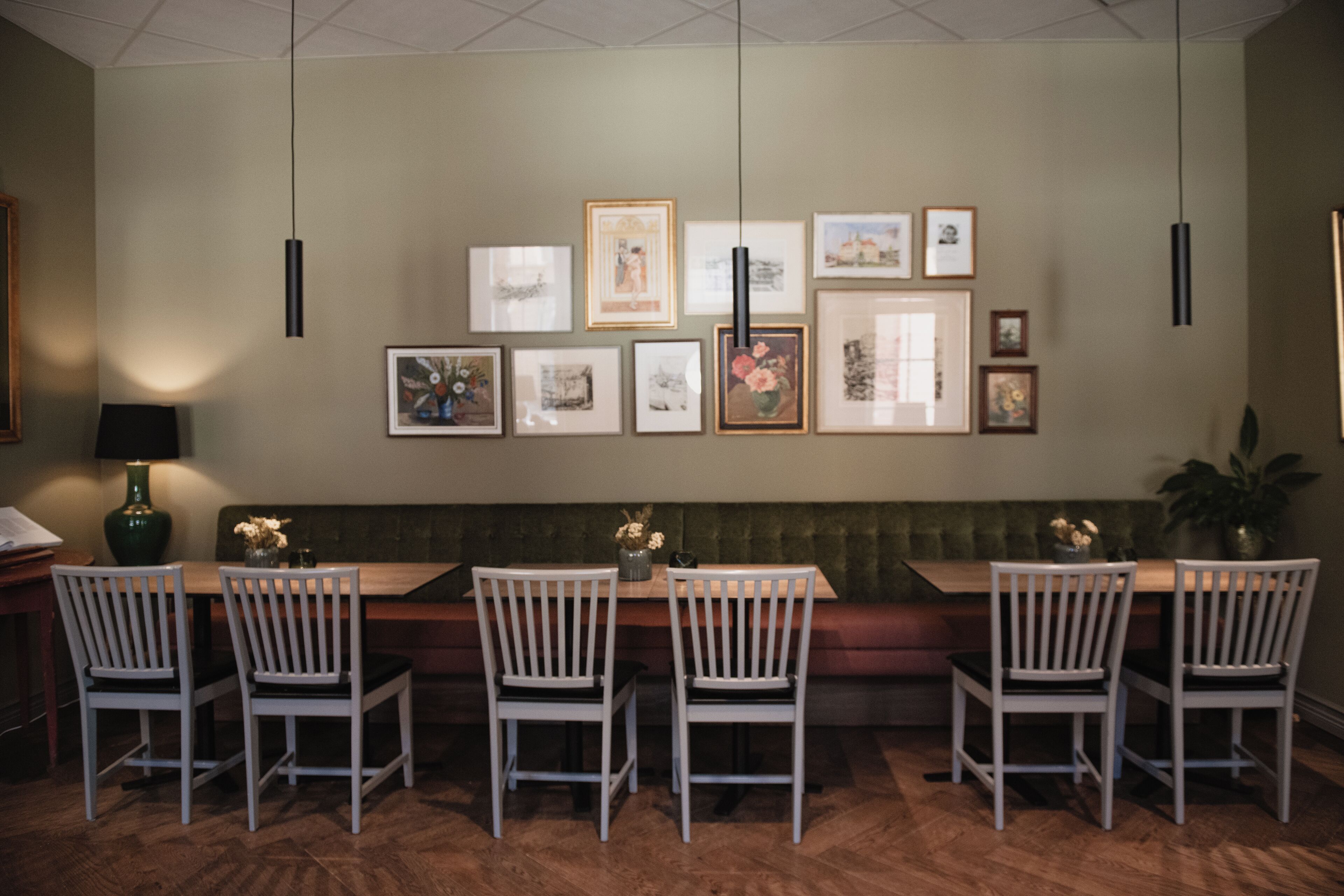 Un comedor con una mesa de madera, sillas blancas y un banco acolchado. Hay cuadros en la pared y un jarrón verde con flores sobre la mesa.