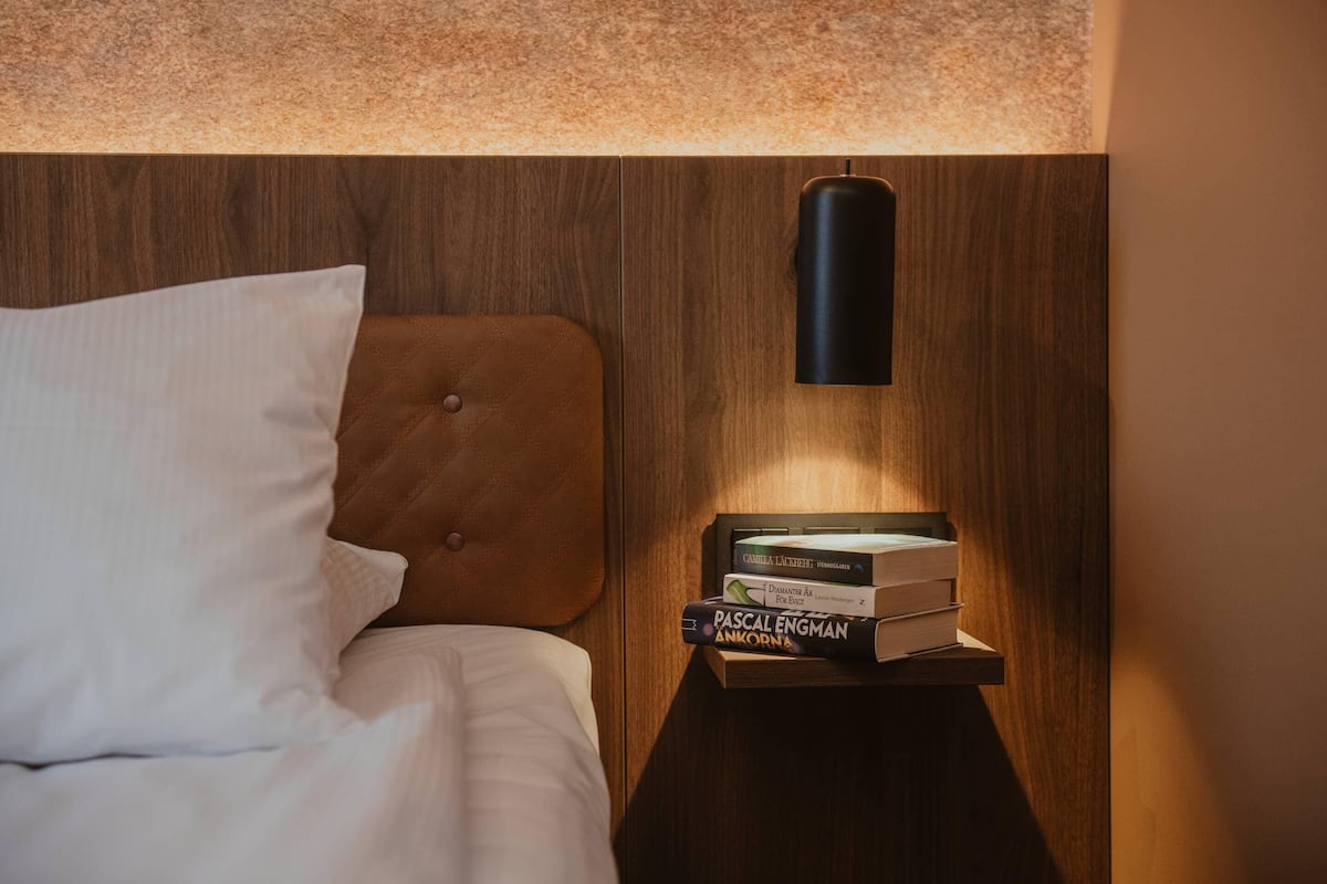 A neatly made bed with a pillow and a headboard, a bedside table with a stack of books, and a wall-mounted lamp.