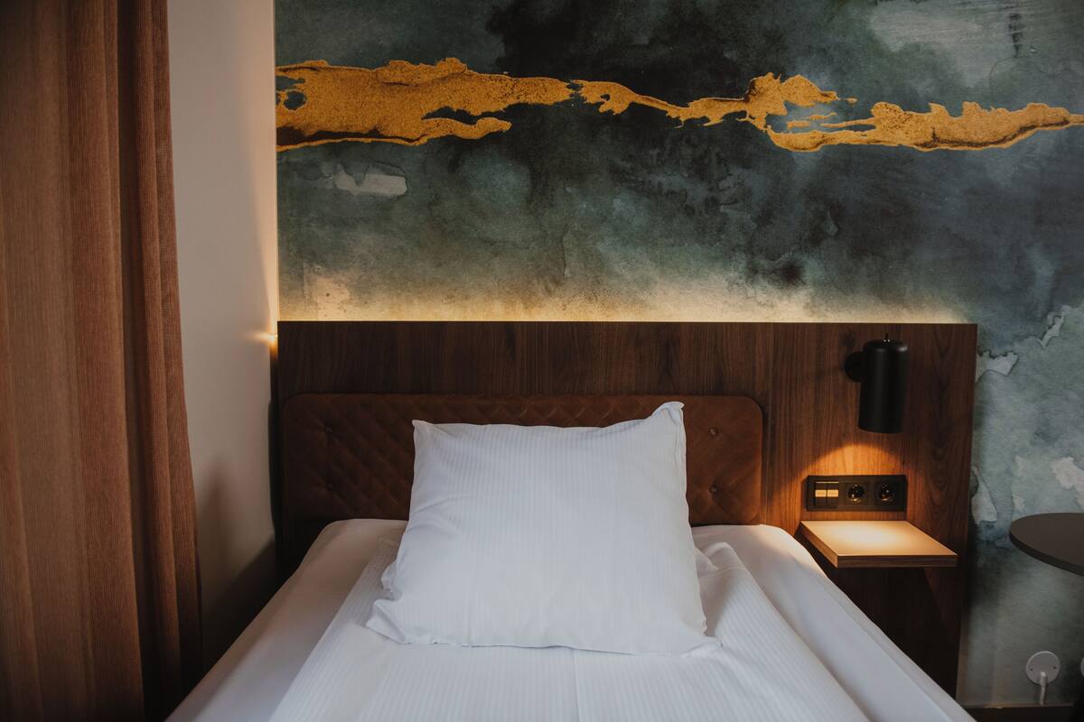 A neatly made bed with a white pillow and a wooden headboard. There is a small bedside table with a lamp and a power outlet. The wall behind the bed features a textured, abstract mural with a mix of dark and light colors.