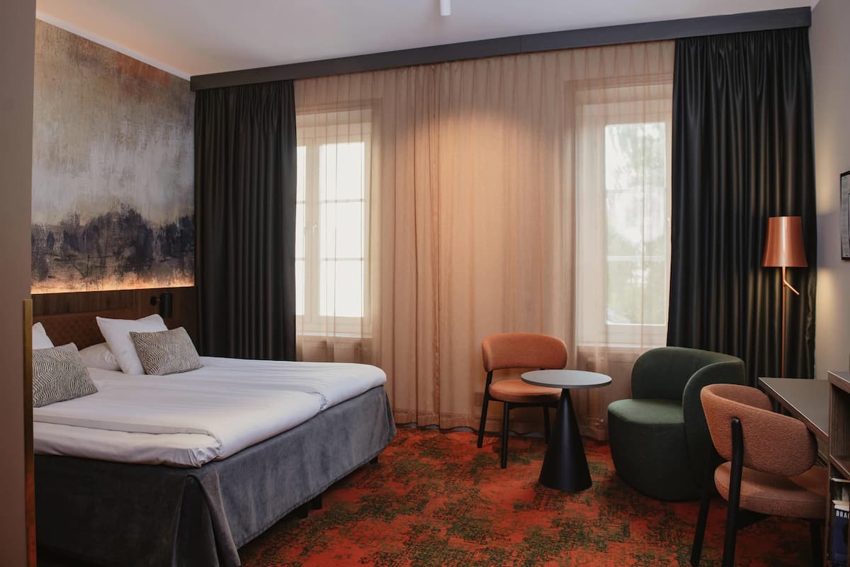 A hotel room with a bed, two chairs, a small table, and a lamp.