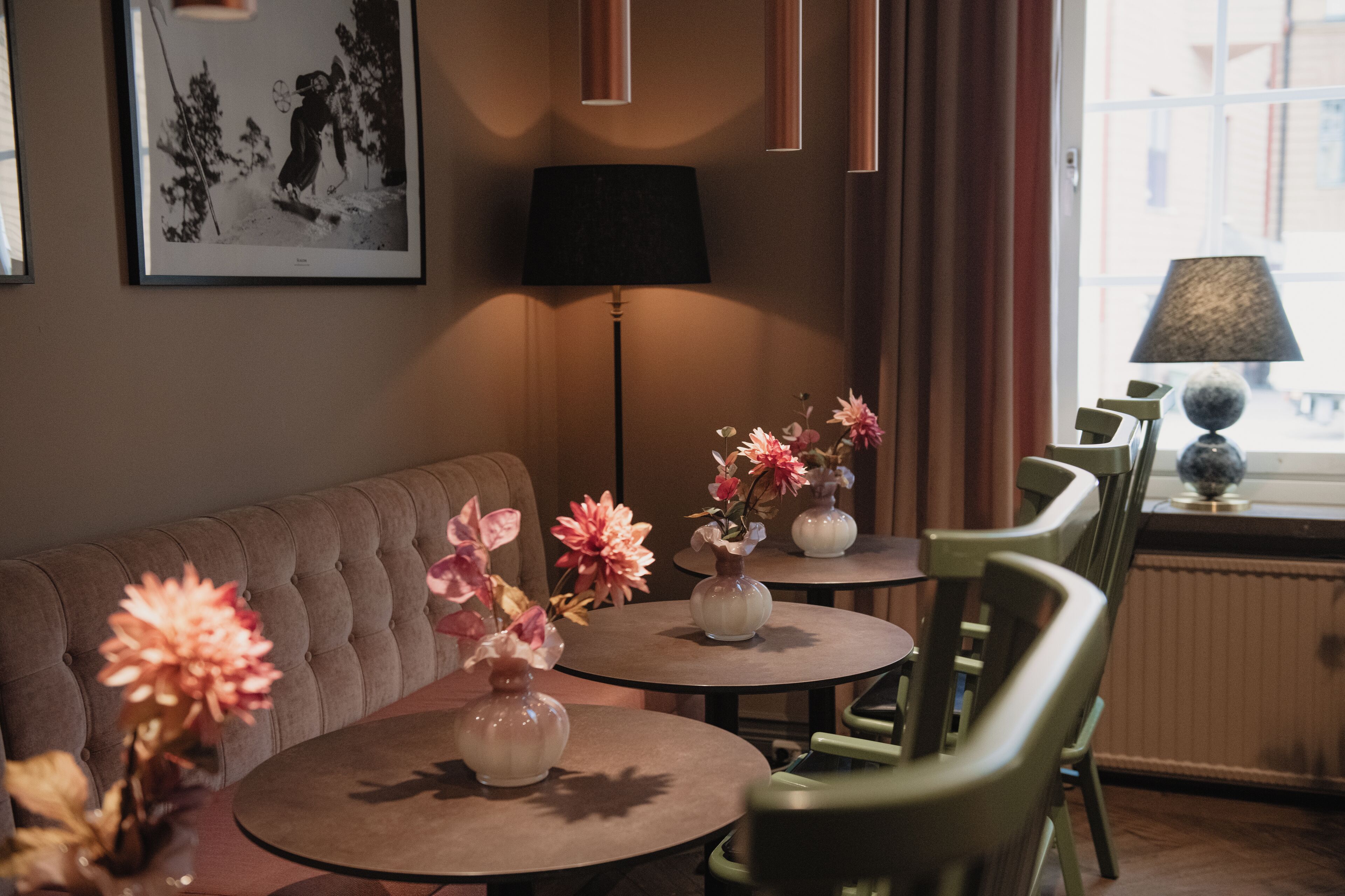 A cozy room with a sofa, two small round tables with vases of flowers, a lamp, and a view of the outside through a window.