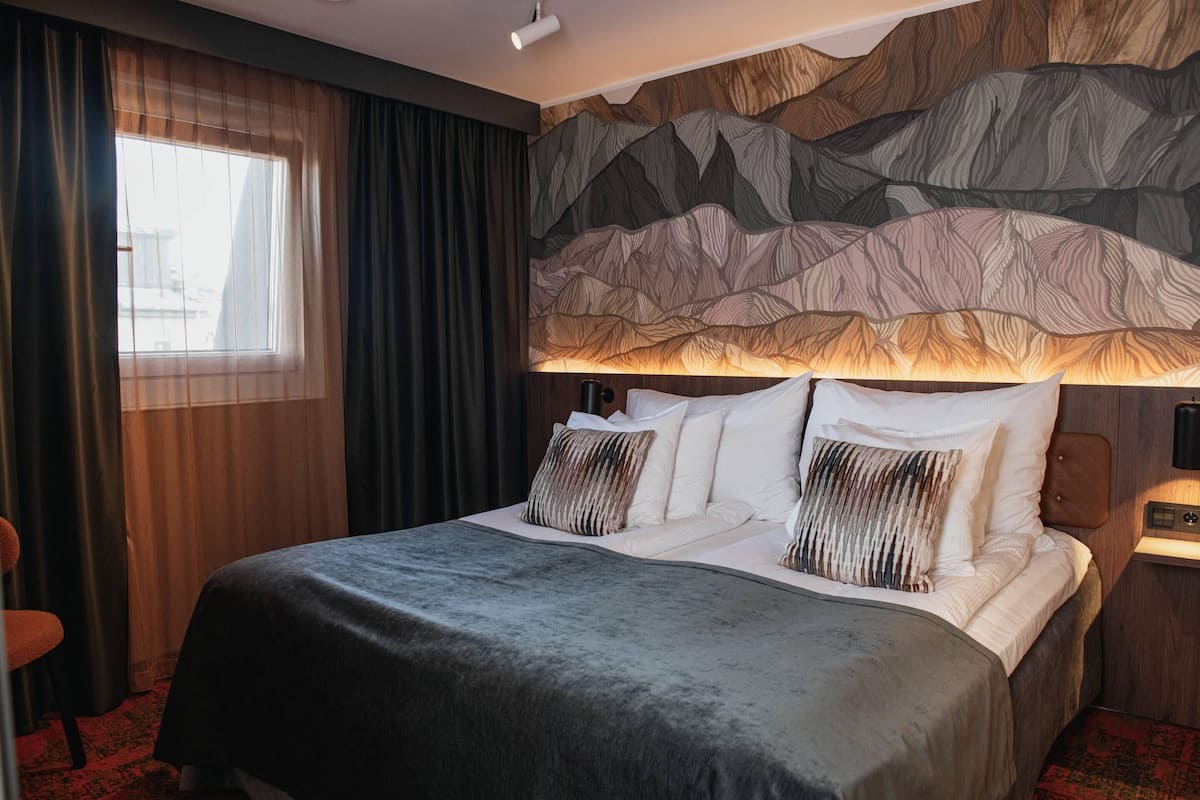 A hotel room with a large bed, a headboard with a mountain design, a window with curtains, and a nightstand with a lamp.