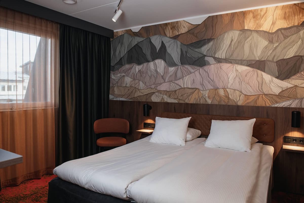 A hotel room with a bed, a chair, a desk, and a wall with a mountain pattern.