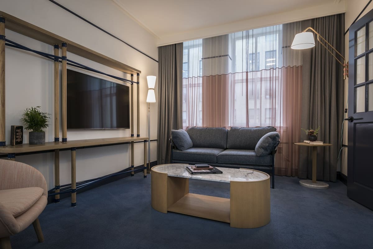 Una habitación de hotel moderna con un sofá gris, una mesa de centro de madera, un televisor montado en un marco de madera y un amplio ventanal con cortinas.