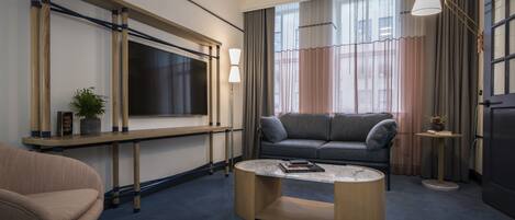 A modern hotel room with a grey sofa, a wooden coffee table, a TV mounted on a wooden frame, and a large window with curtains.