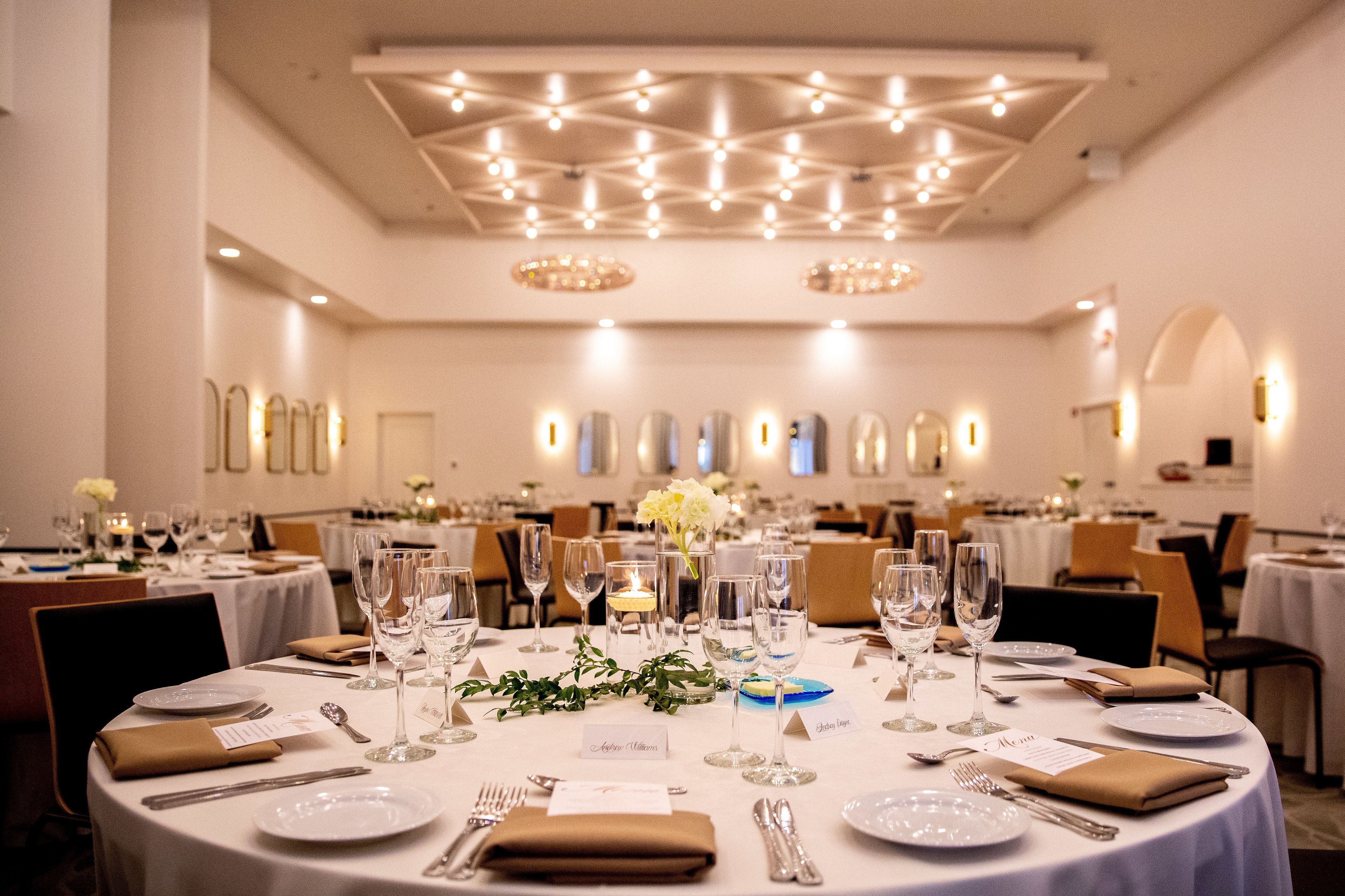 Ein Festsaal mit runden Tischen, gedeckt für eine festliche Veranstaltung, mit weißen Tischdecken, Gläsern und floralen Mittelpunkten.