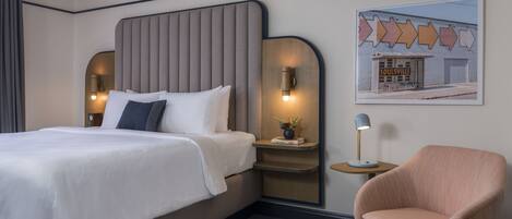 A hotel room with a large bed, a nightstand with a lamp, a chair, and a wall-mounted artwork.