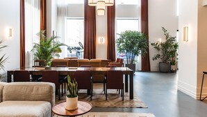Uma área de refeições moderna com mesa de madeira, cadeiras vermelhas e um lustre central. Há plantas em vasos e uma grande janela com cortinas.
