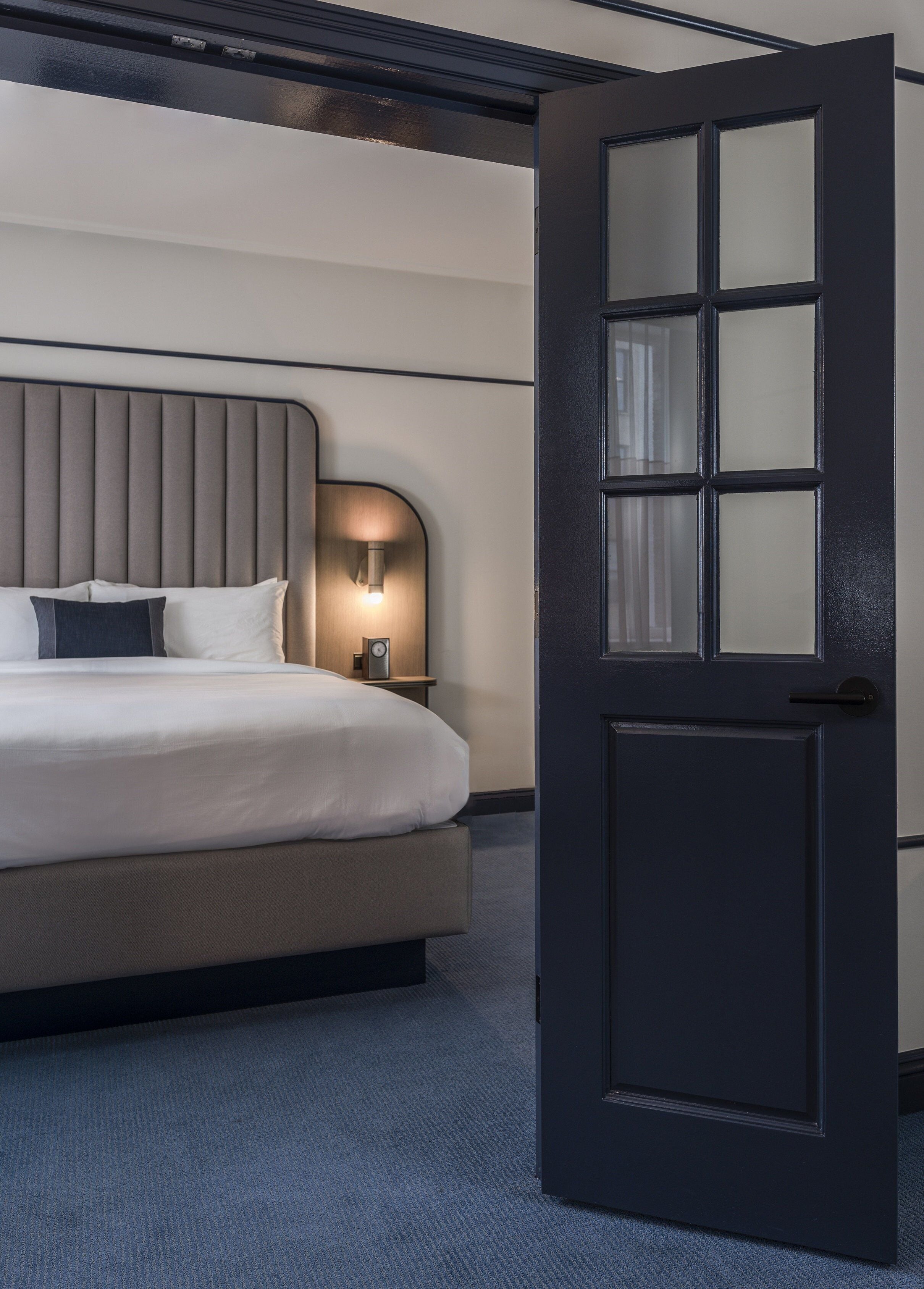 A hotel room with a bed, a nightstand, a wall-mounted light, and a door.