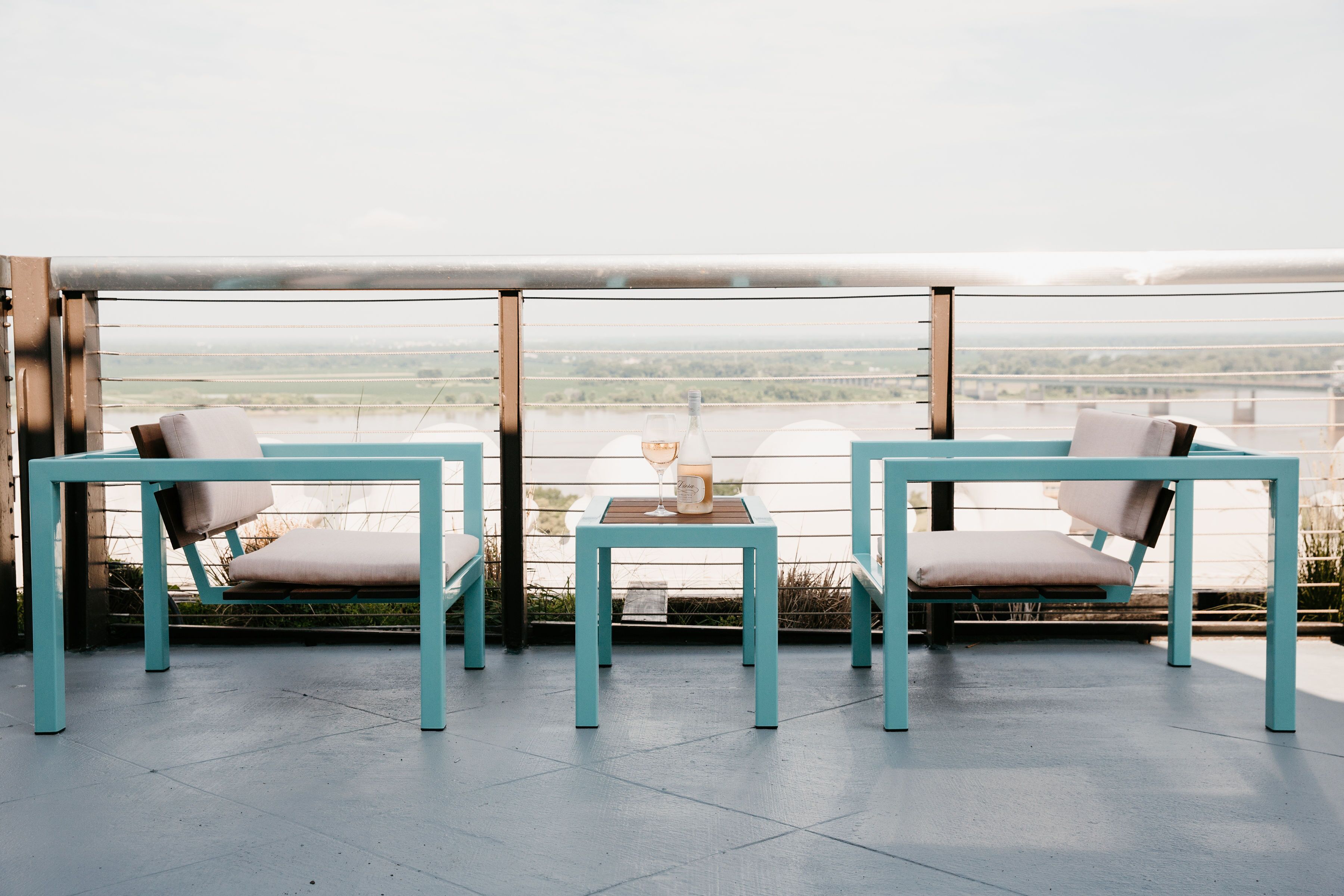 A terrace with two turquoise tables and chairs, a bottle of wine, and a view of a bridge and landscape.