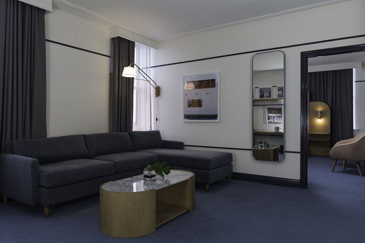 A modern hotel room with a grey sectional sofa, a round coffee table, a large mirror, and a small plant.