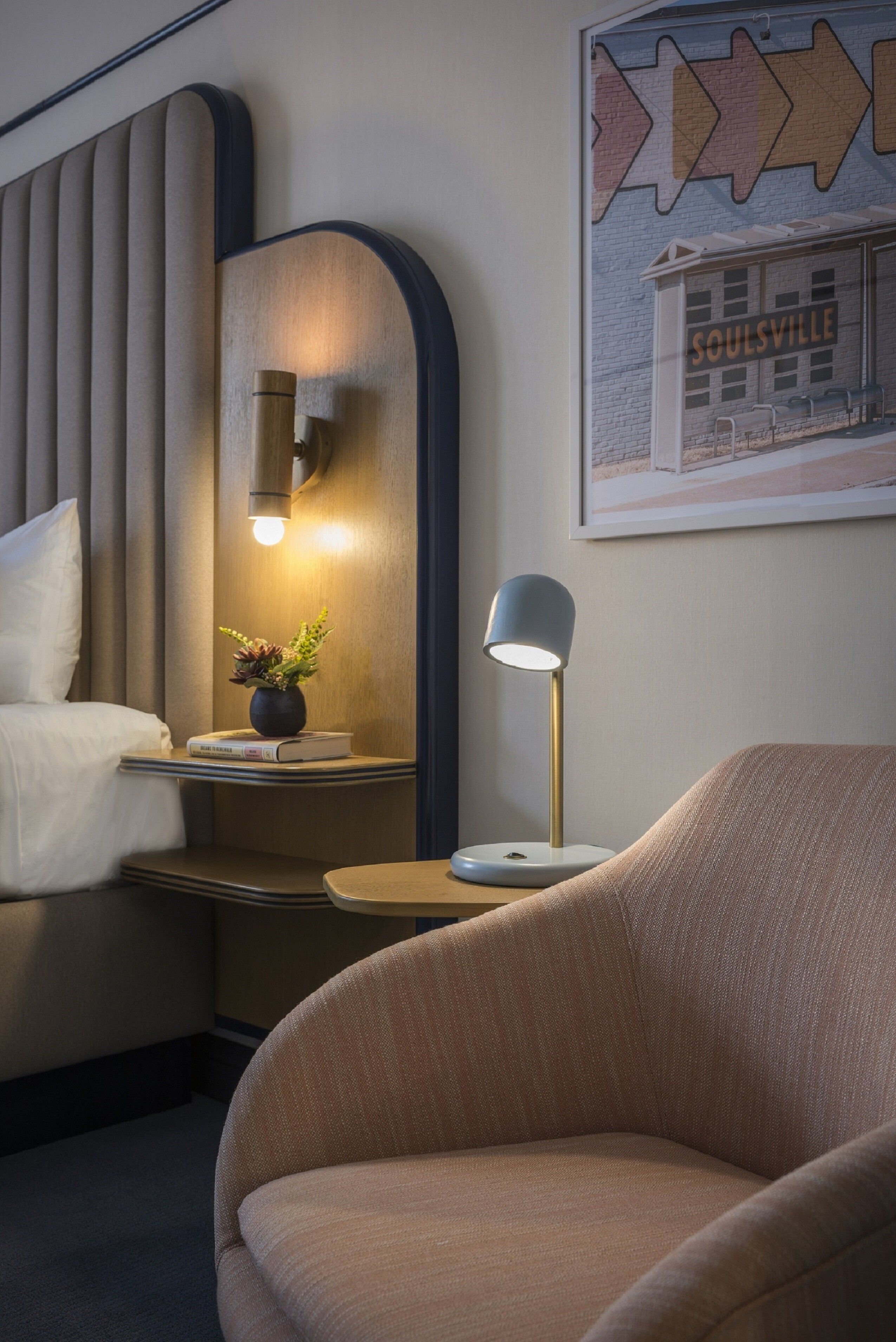 A hotel room with a bed, bedside table with a lamp, a small plant, and a framed picture on the wall.