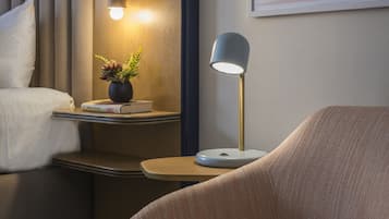 A hotel room with a bed, bedside table with a lamp, a small plant, and a framed picture on the wall.
