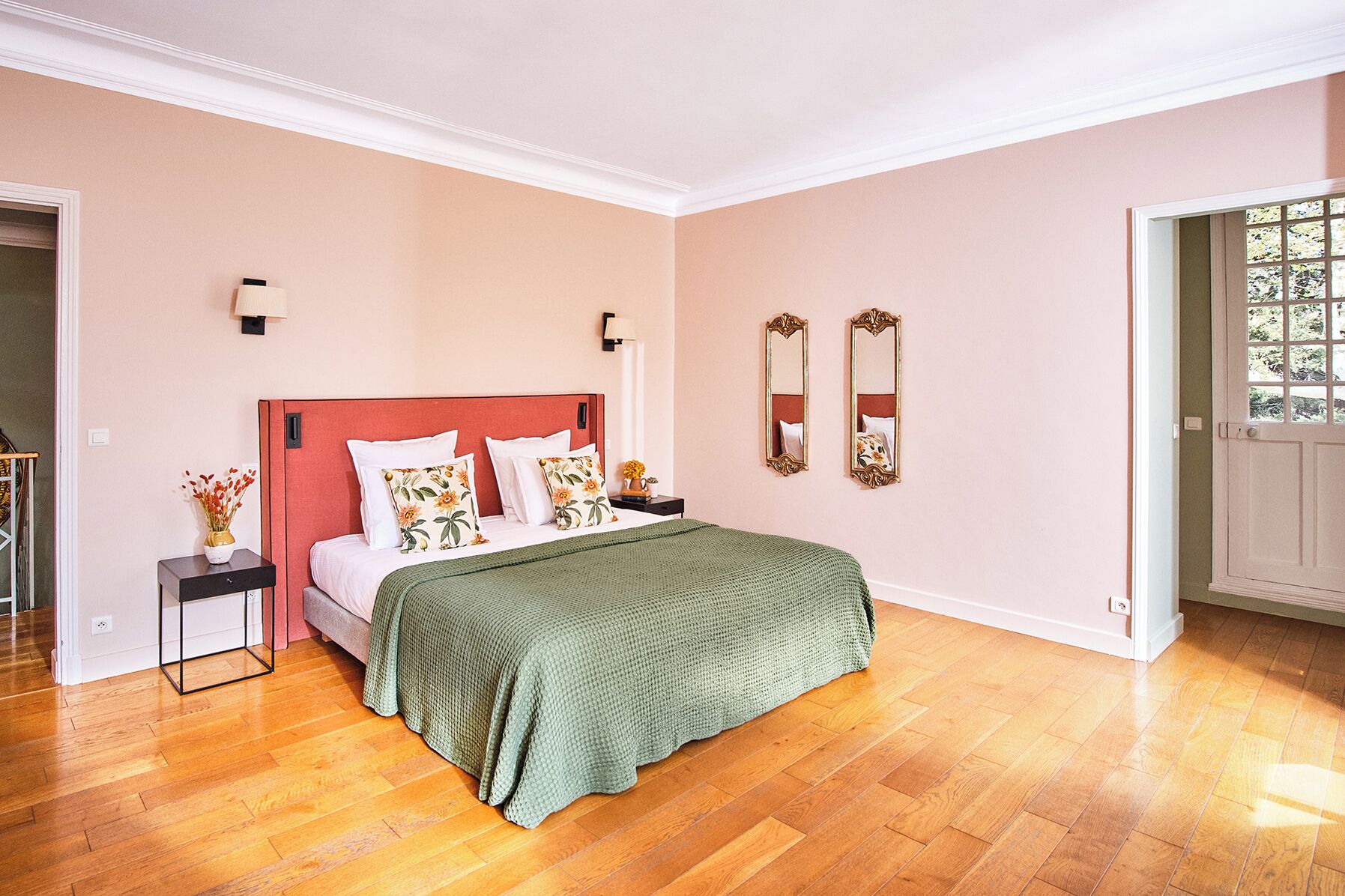 A bedroom with a wooden floor, a bed with a green blanket, two nightstands, a mirror, and a door leading to a balcony.