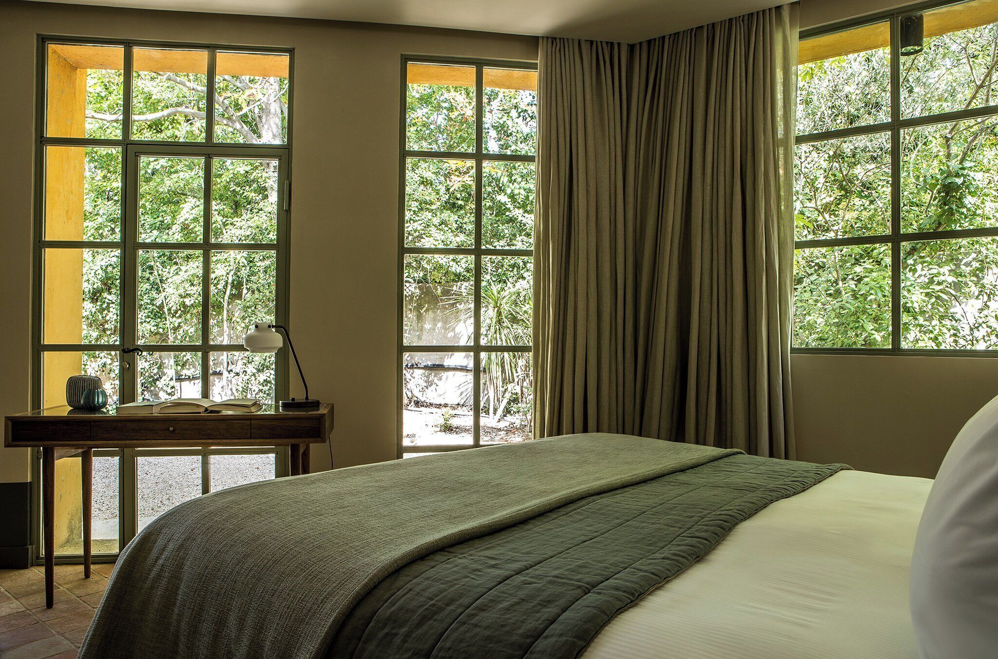 A hotel room with a large bed, a desk with a lamp, and a view of greenery through large windows.
