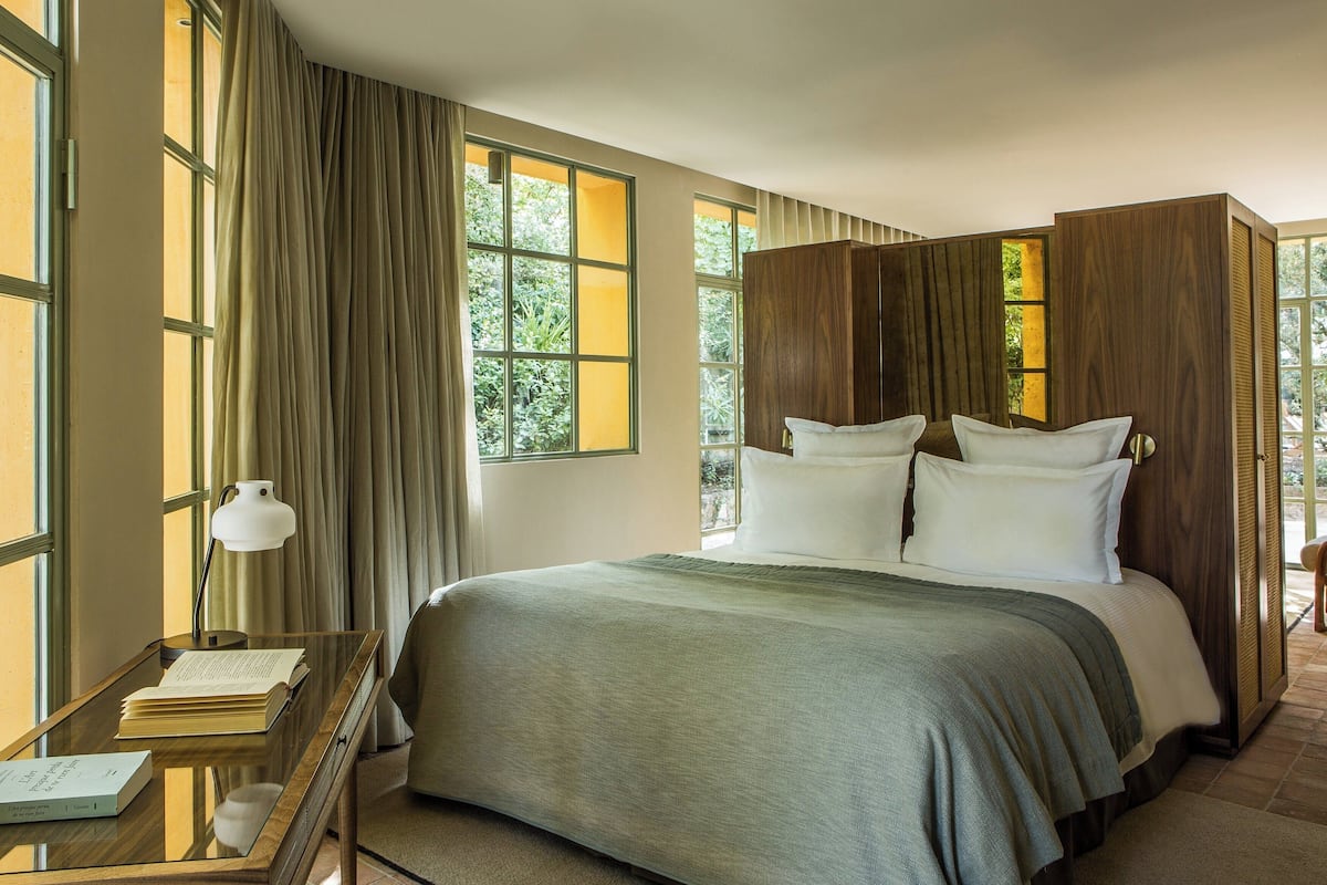 A hotel room with a large bed, a desk with a lamp, and a view of greenery through the windows.