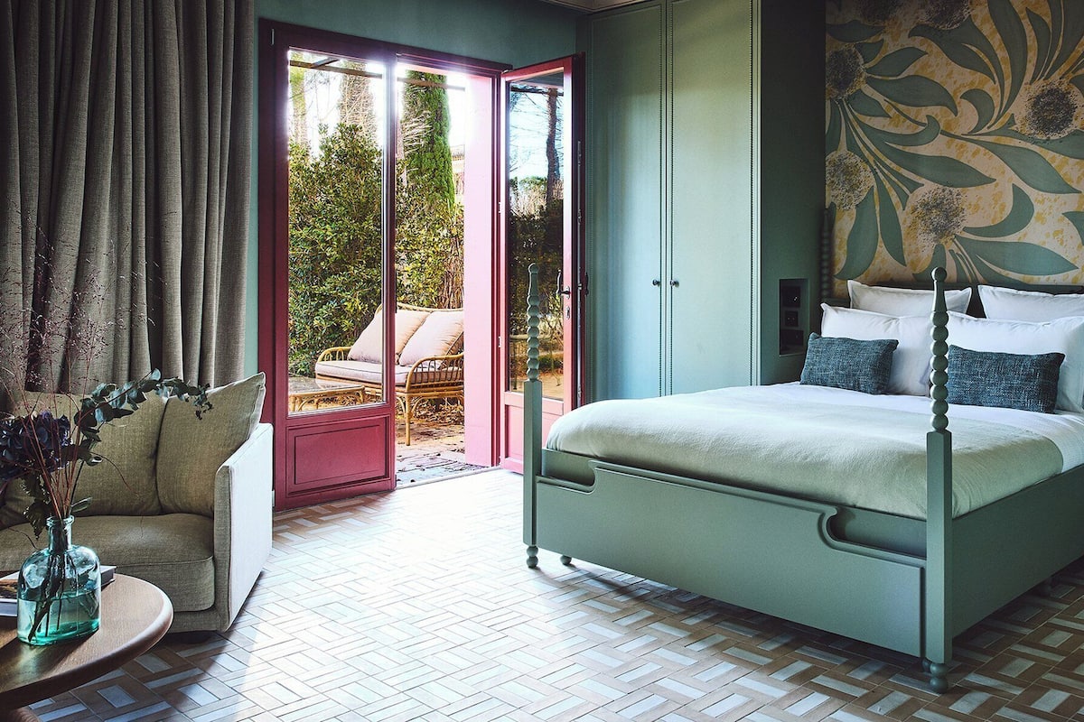 A bedroom with a four-poster bed, a sofa, a table with a vase, and a view of a garden through a glass door.