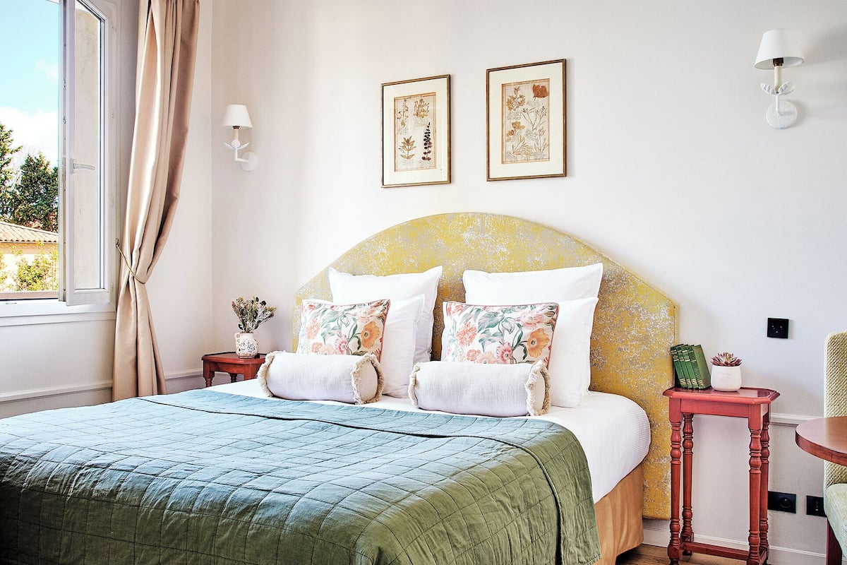 A bedroom with a bed, two wall-mounted lamps, a window with curtains, and two framed botanical prints on the wall.
