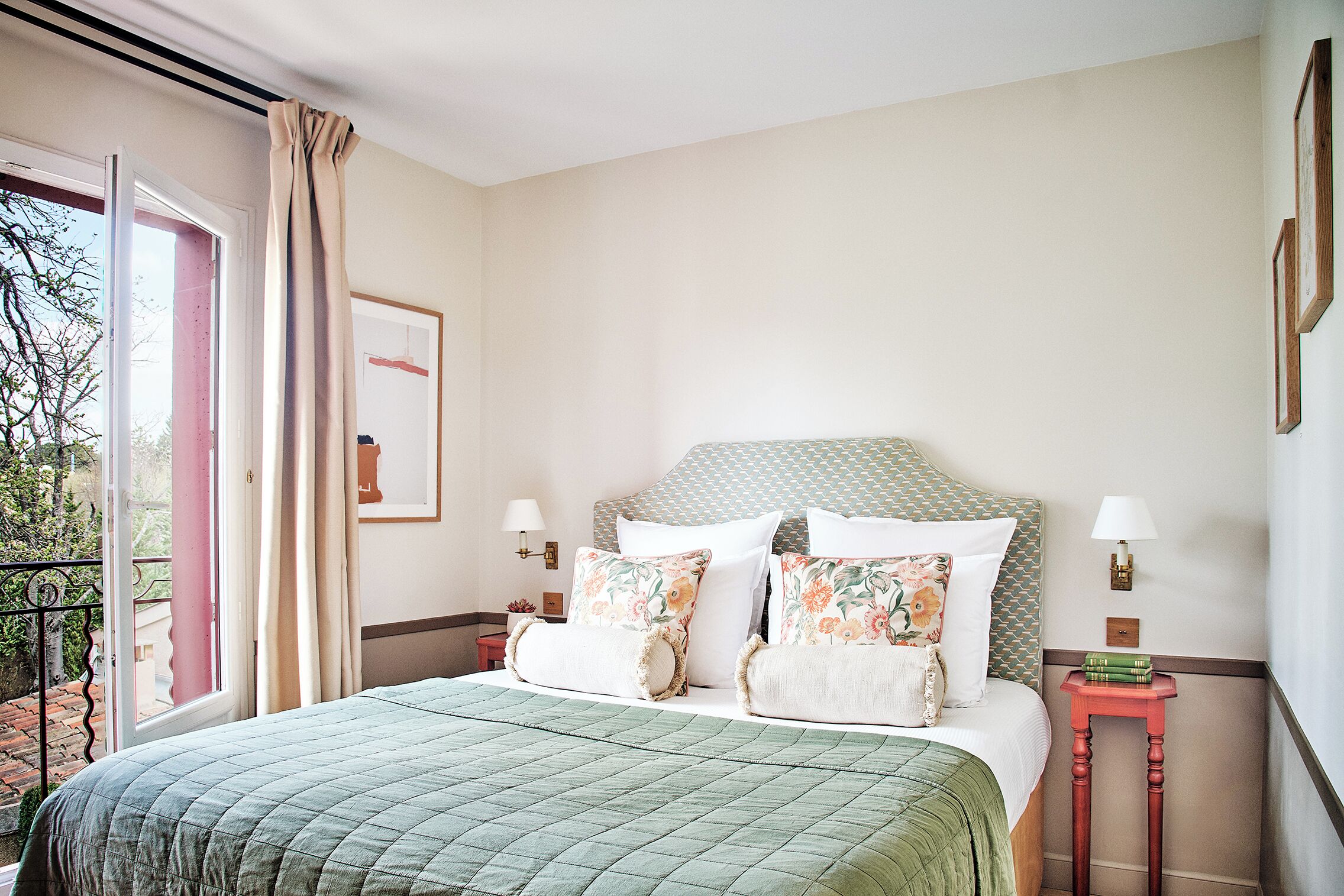 A neatly made bed with a green quilt, a headboard with a patterned design, and a small bedside table with a lamp and books.