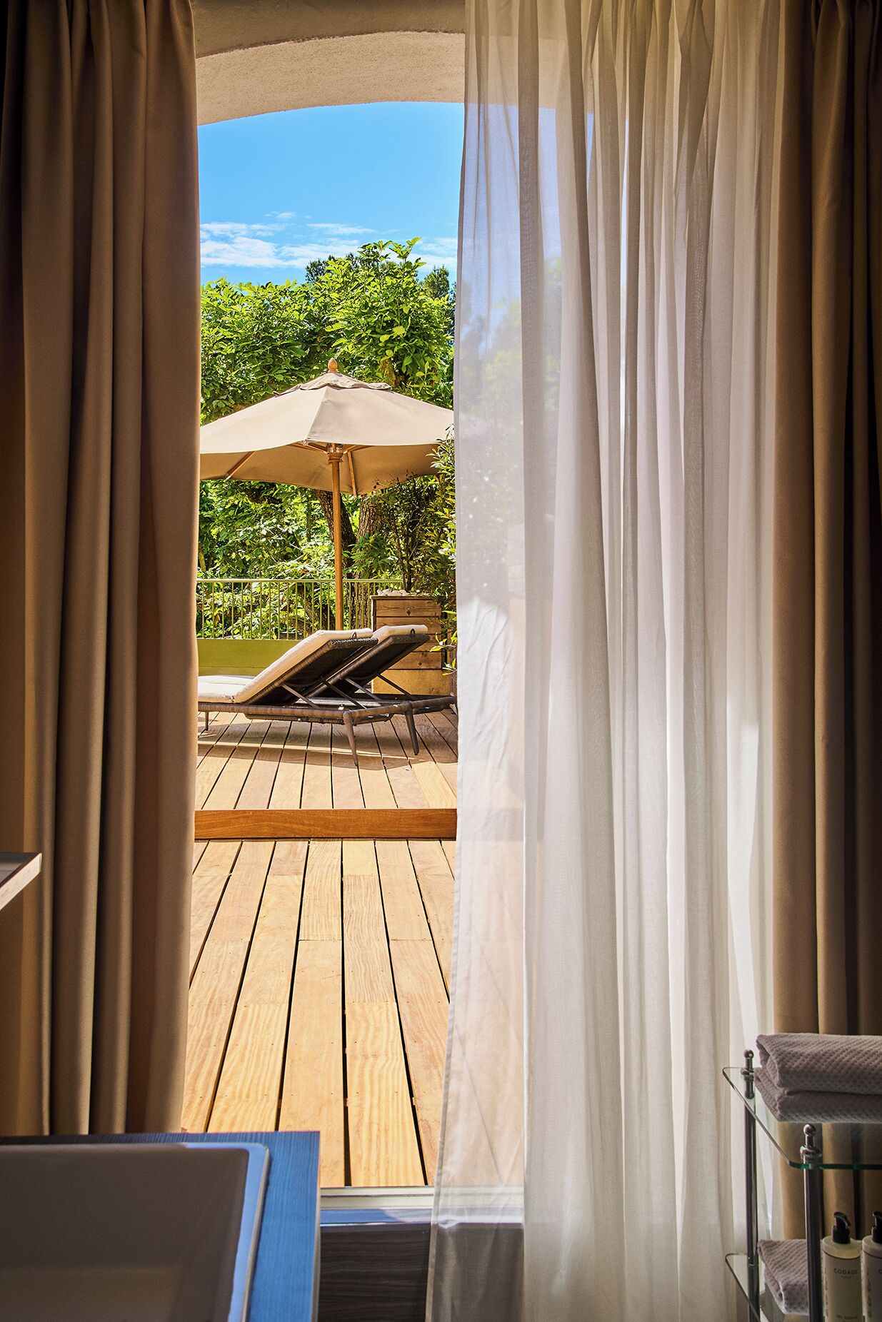 Una terraza de madera con una silla de descanso y una sombrilla, una mesa de madera y un estante de vidrio con toallas y artículos de aseo.