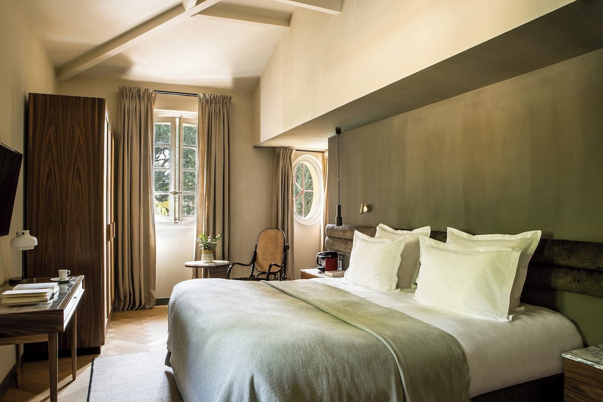 A hotel room with a large bed, a desk with a lamp, a chair, and a view of the outdoors through a window.