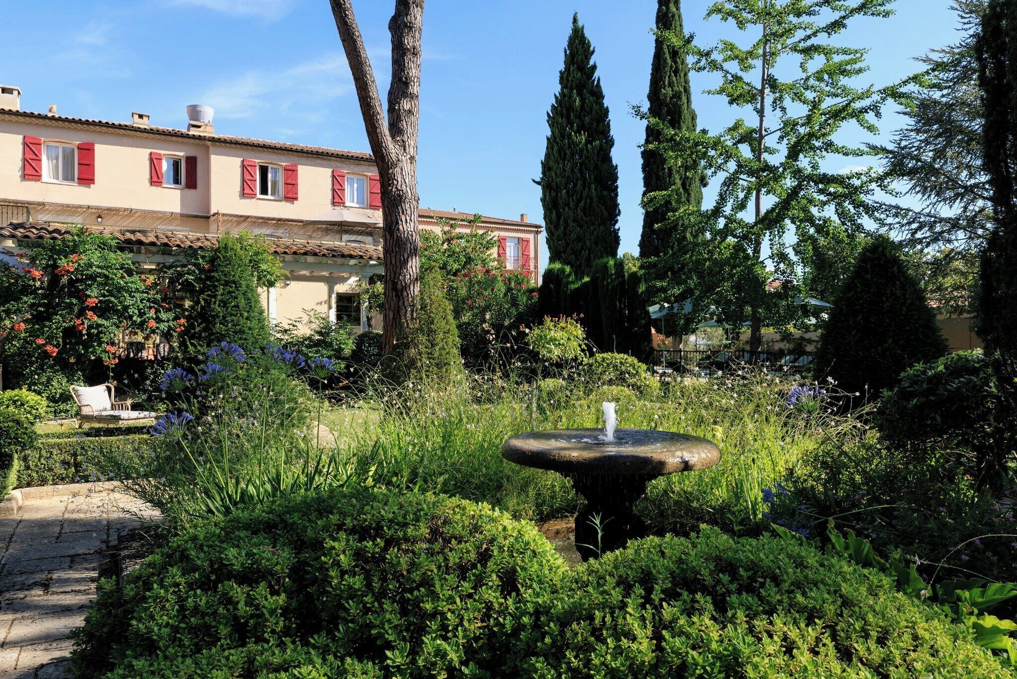A garden with a fountain, lush greenery, and a building with red shutters in the background.