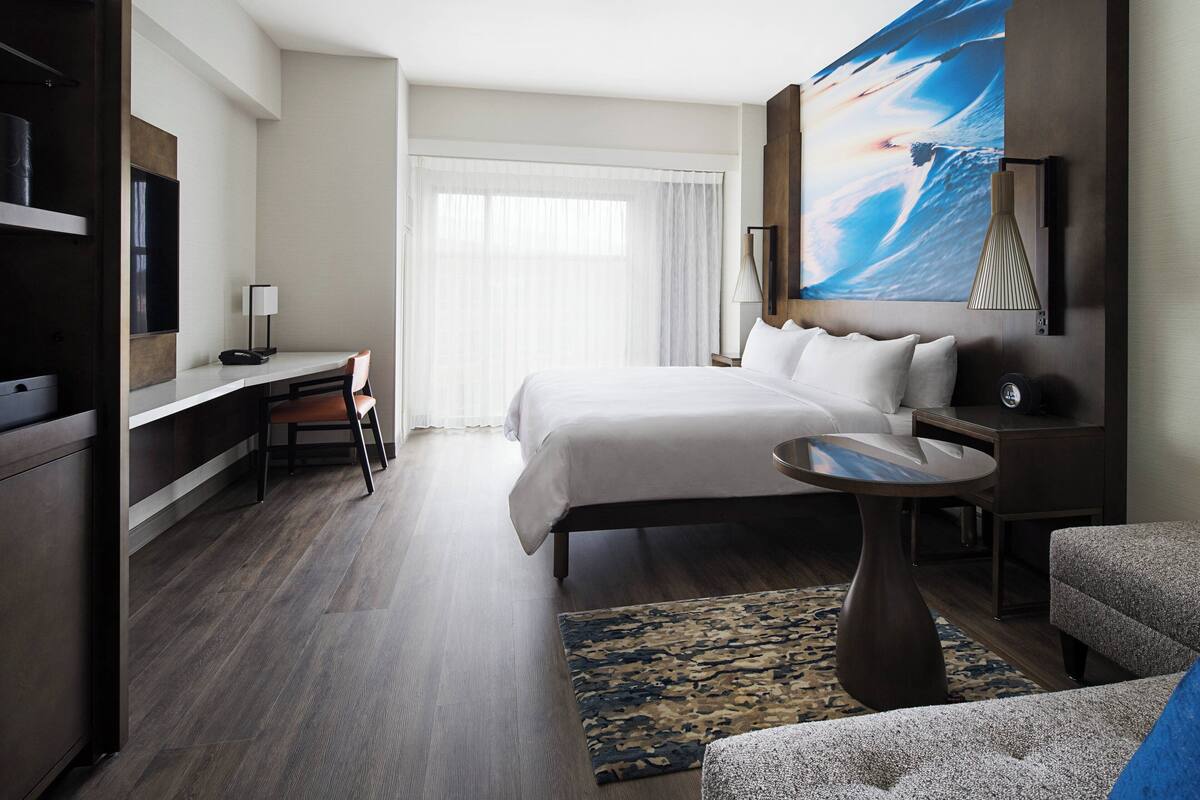A hotel room with a large bed, a desk with a chair, a TV, and a painting on the wall.