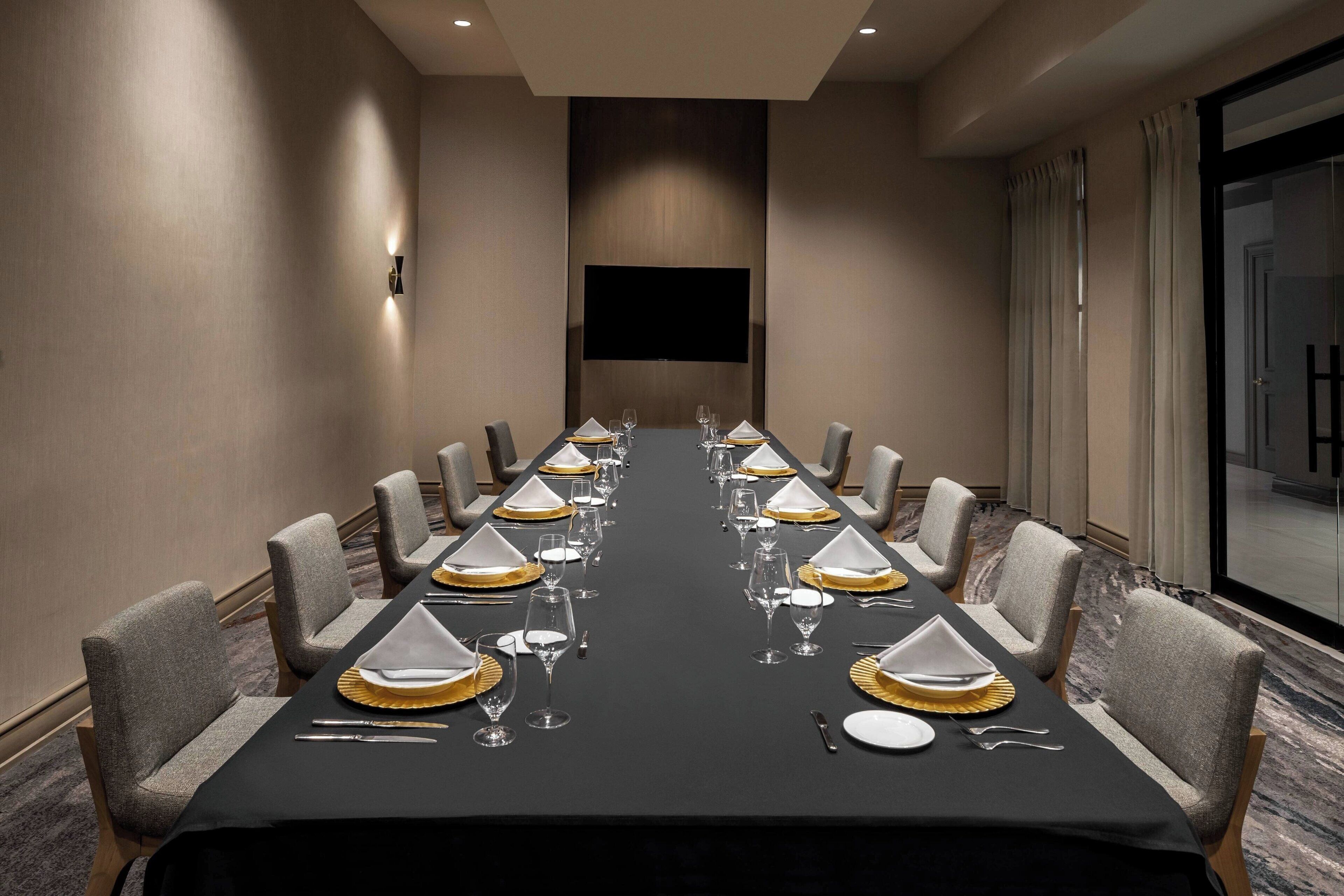 Un salón de conferencias con una larga mesa dispuesta para un evento formal, equipado con sillas, vajilla y un televisor de pantalla plana.