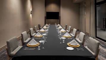 Un salón de conferencias con una larga mesa dispuesta para un evento formal, equipado con sillas, vajilla y un televisor de pantalla plana.