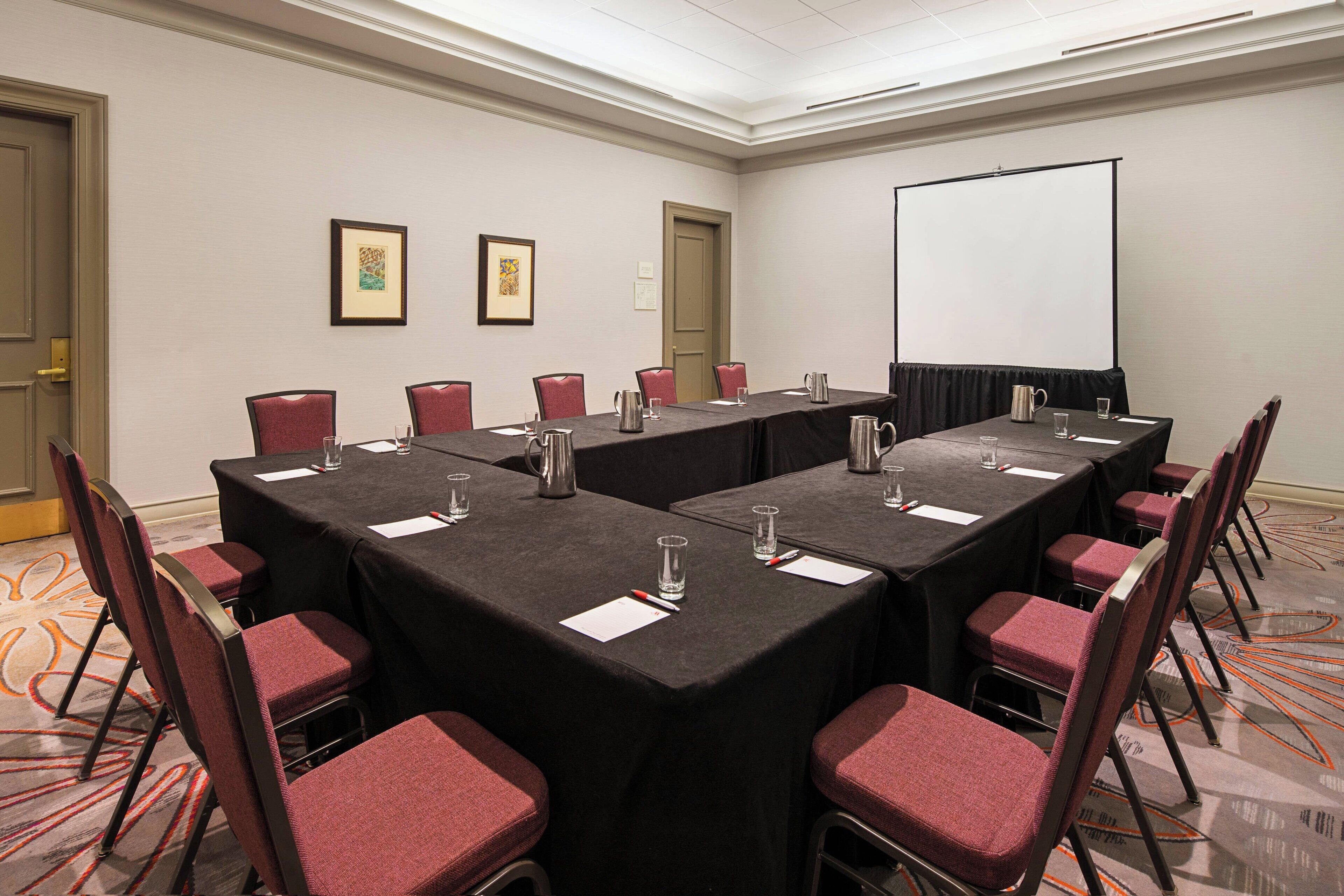Ein Konferenzraum mit einem langen Tisch, Stühlen, einer Projektionsleinwand und gerahmten Bildern an der Wand.