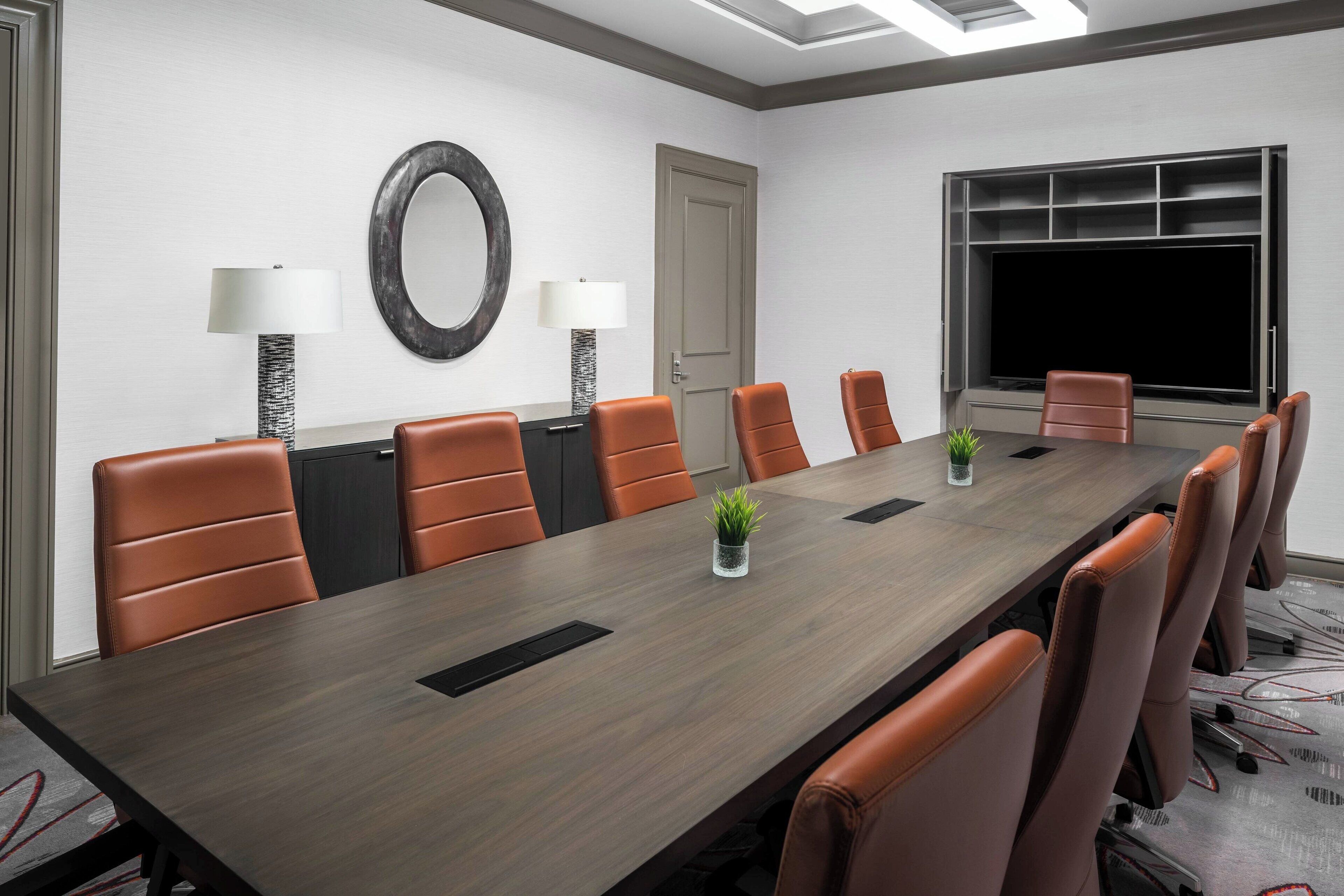 A conference room with a long table, brown leather chairs, two lamps, a potted plant, and a large window.